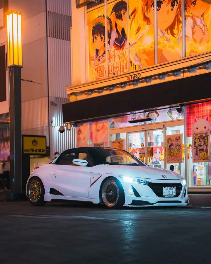 Una Honda S660 bianca con i fari accesi, davanti a un negozio illuminato di Tokyo