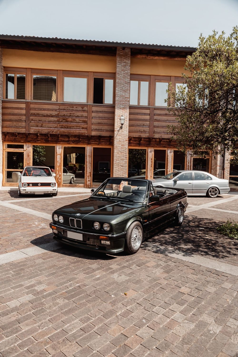 Una BMW E30 cabrio nera ferma su un selciato davanti a un edificio in legno