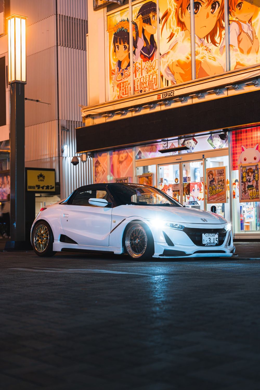 Una Honda S660 bianca di notte, con i fari accesi davanti a una vetrina illuminata